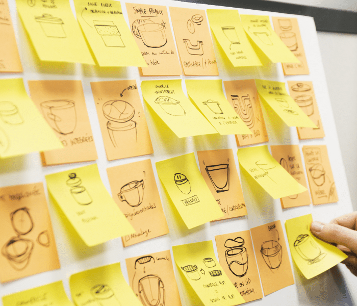 Un tableau rempli de post-its jaunes avec divers croquis et annotations pour tester une idée de produit ou de service. Une main tient un post-it, suggérant une session de brainstorming en cours.