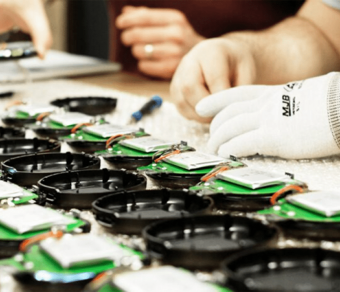 Des mains gantées assemblent des composants électroniques verts et noirs sur une table, représentant une ligne de production lors de la phase d'industrialisation.